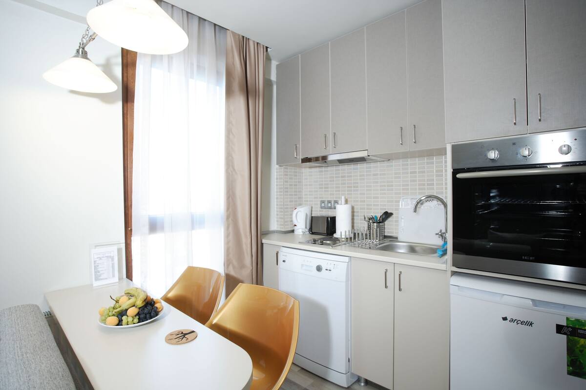 A modern kitchen with a dishwasher, oven, and microwave. There is a dining table with a fruit bowl and chairs.