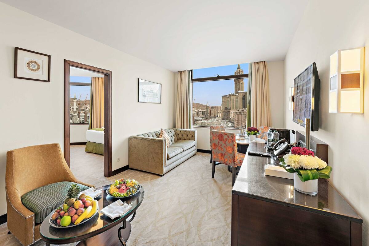 A hotel room with a sofa, armchair, dining table with fruit, a desk with a phone, and a large window with a city view.