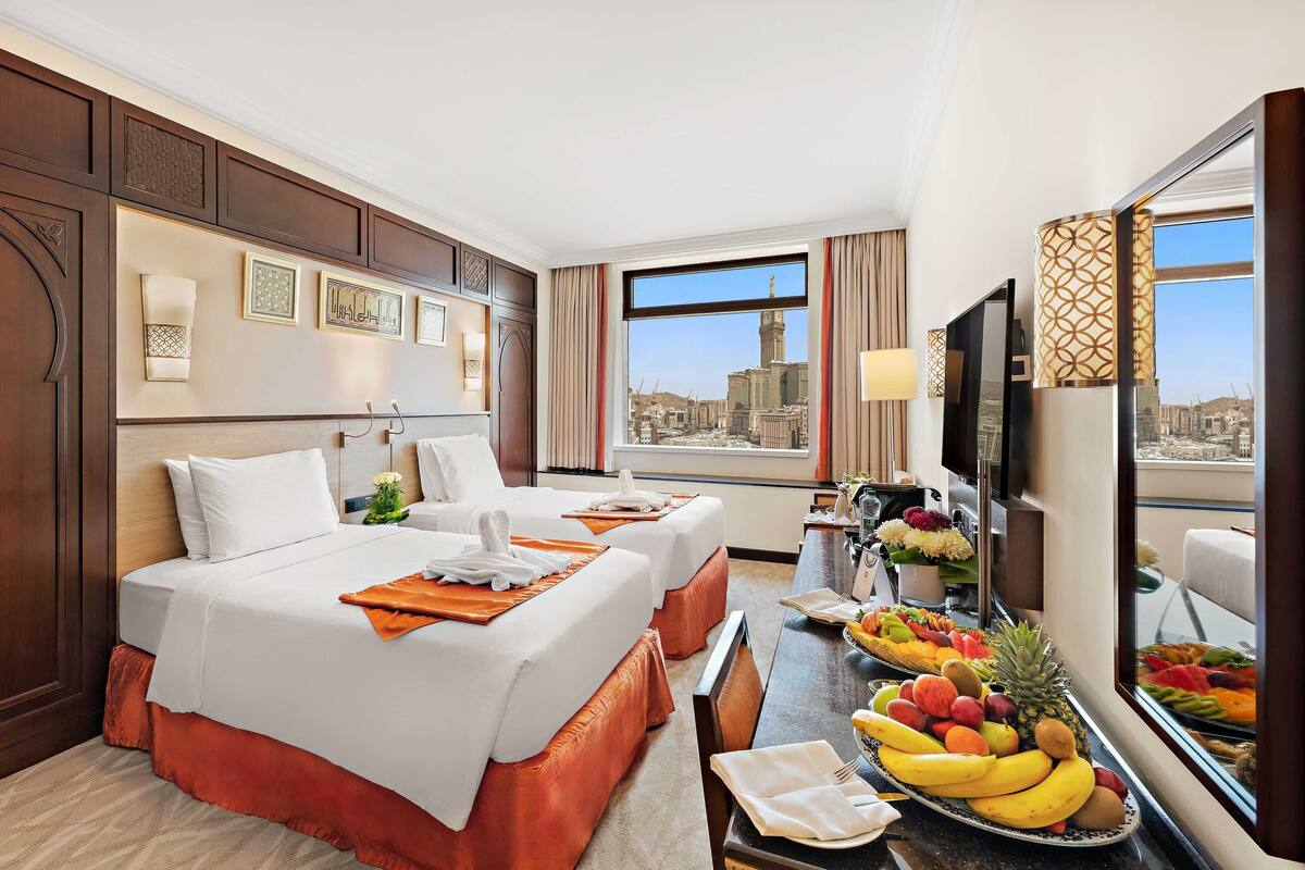 A hotel room with two beds, a dining table with fruit, a TV, and a city view.