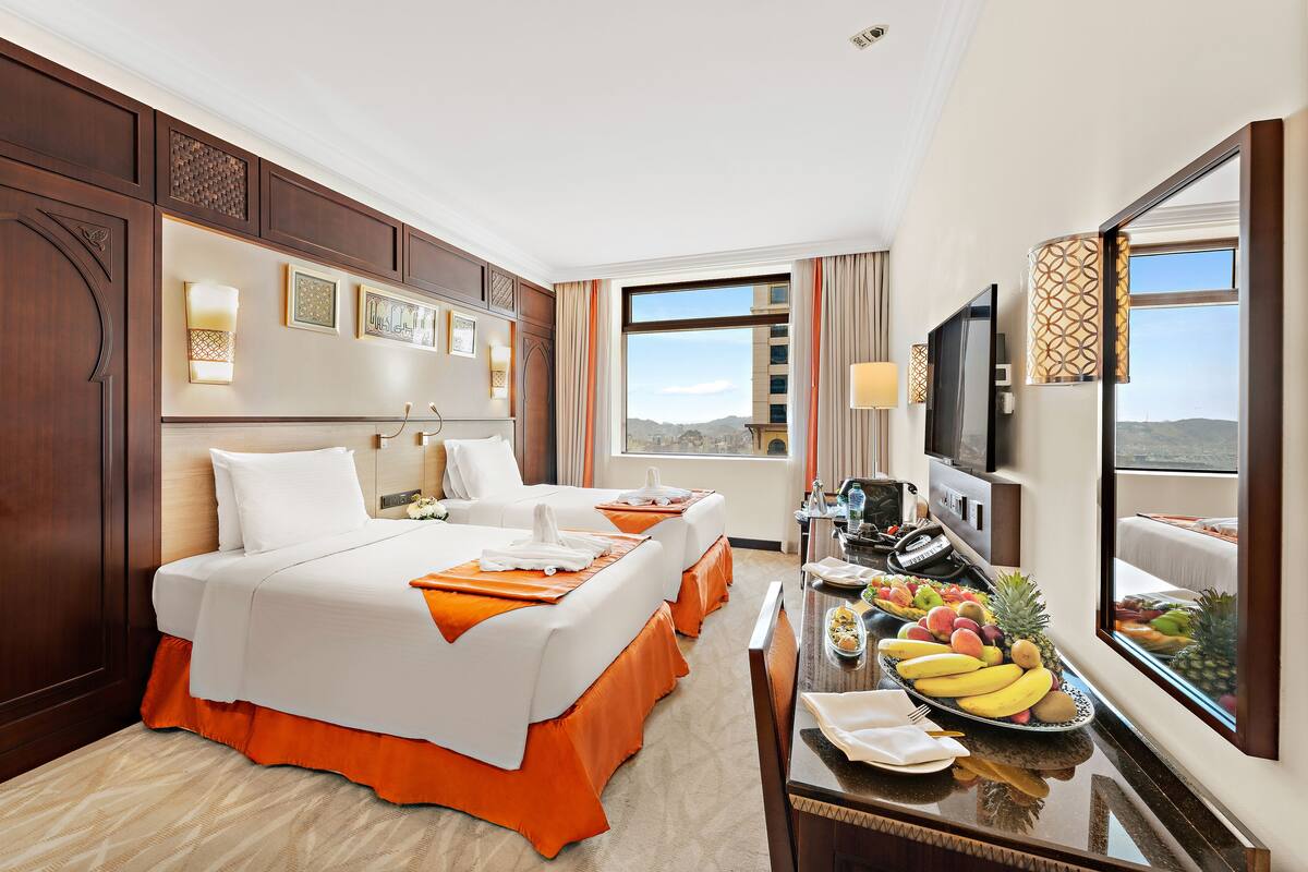 A hotel room with two beds, a dining table with fruit, a TV, and a large window with a city view.