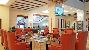 A lobby with red leather chairs, a glass coffee table, and a chandelier. There is a staircase in the background and a TV mounted on the wall.