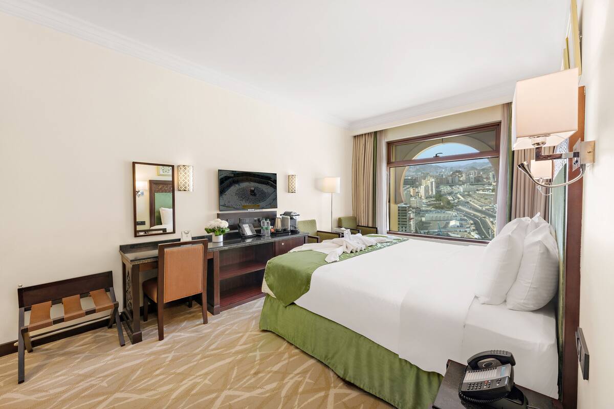 A hotel room with a large bed, a desk with a chair, a small table with a phone, a bench, a TV, and a window with a city view.