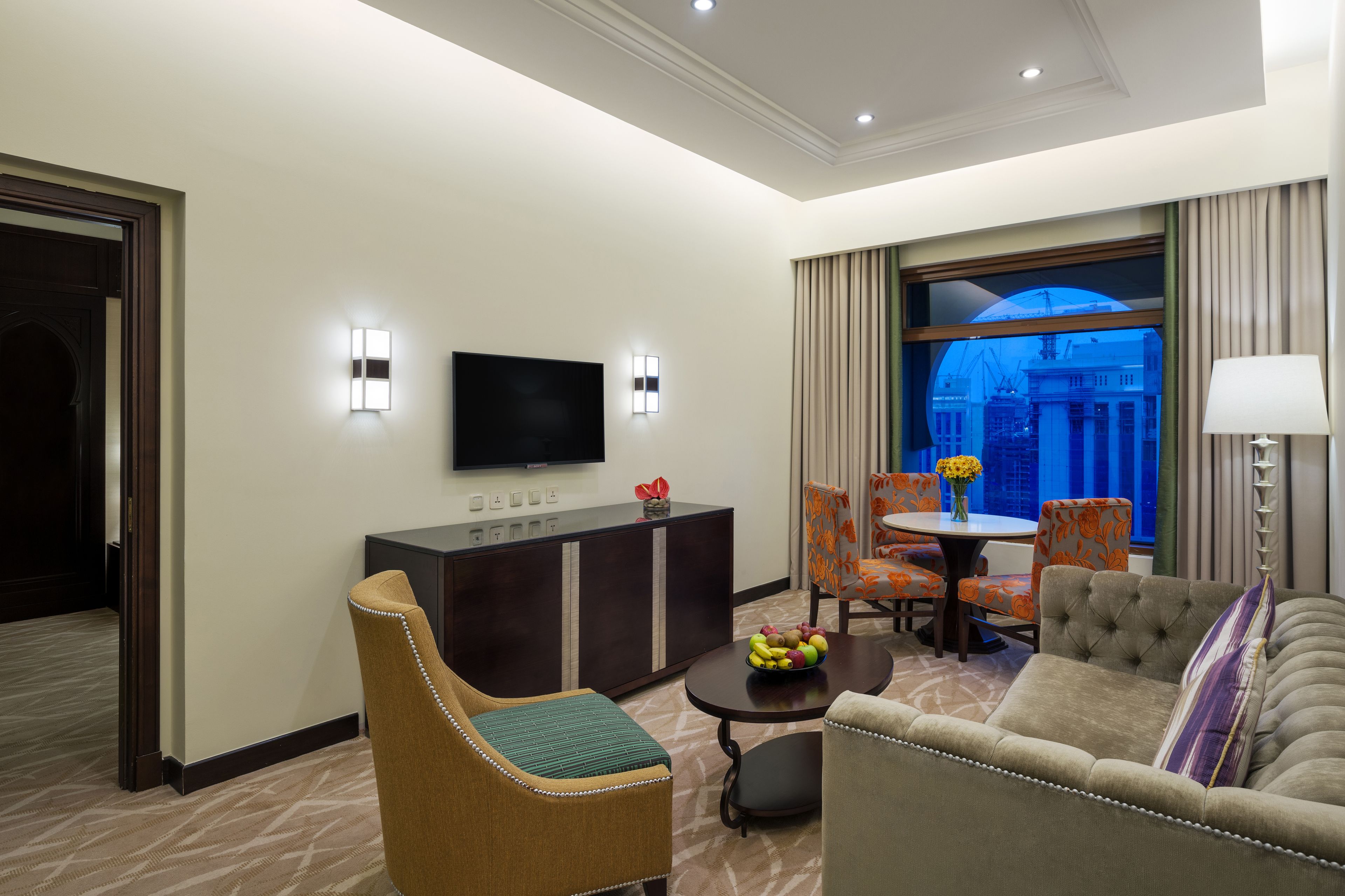 A hotel room with a flat-screen TV, a sofa, a dining table with chairs, and a view of a cityscape through a large window.