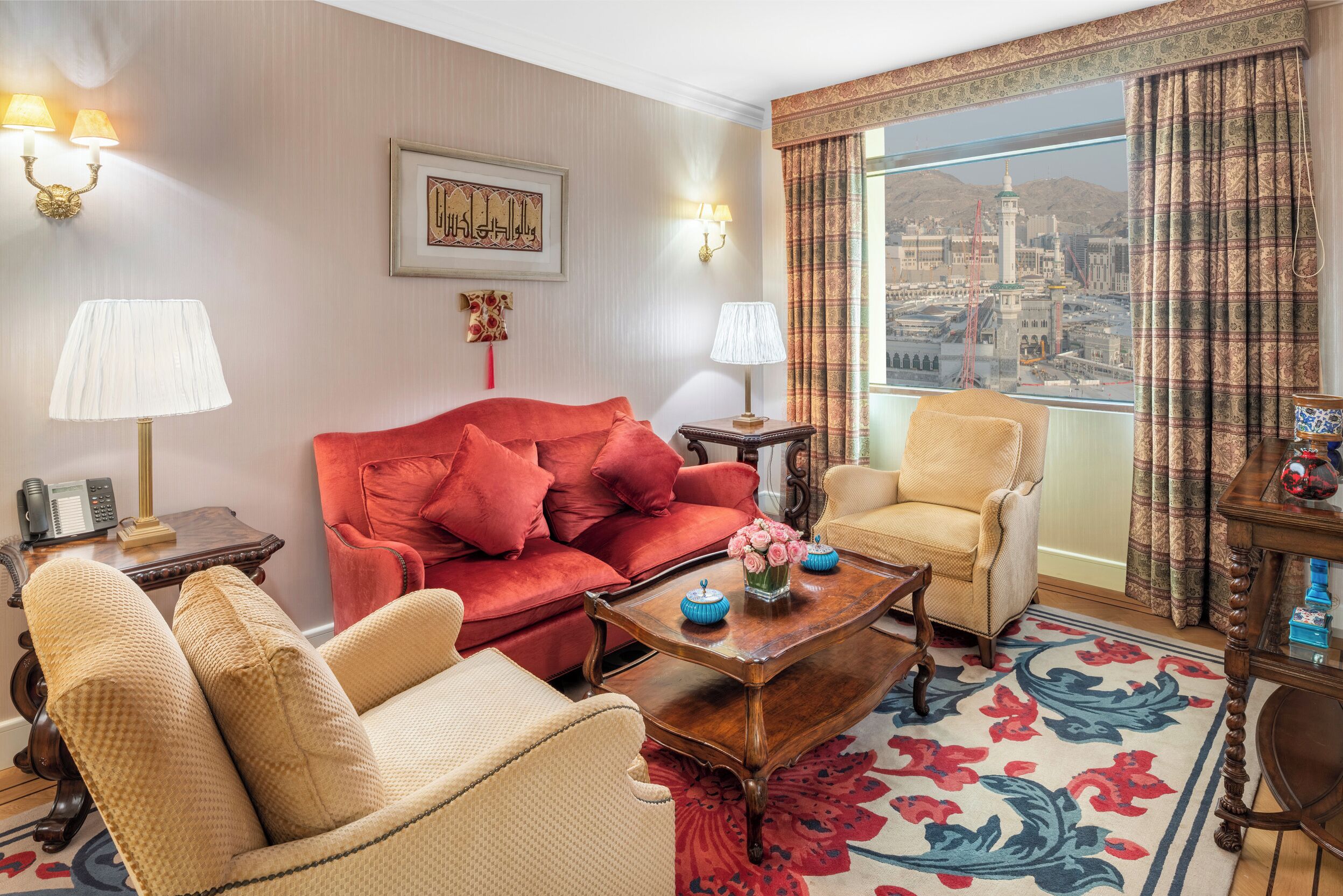A hotel room with a red sofa, beige armchairs, a wooden coffee table, and a large window with a view of a cityscape.