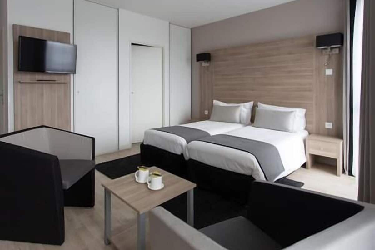 A modern hotel room with a large bed, a TV mounted on the wall, a small table with cups, and a seating area with a chair.