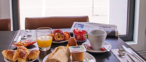 Una mesa de desayuno con croissants, huevos y jugo de naranja, frente a una ventana con vista a un edificio.