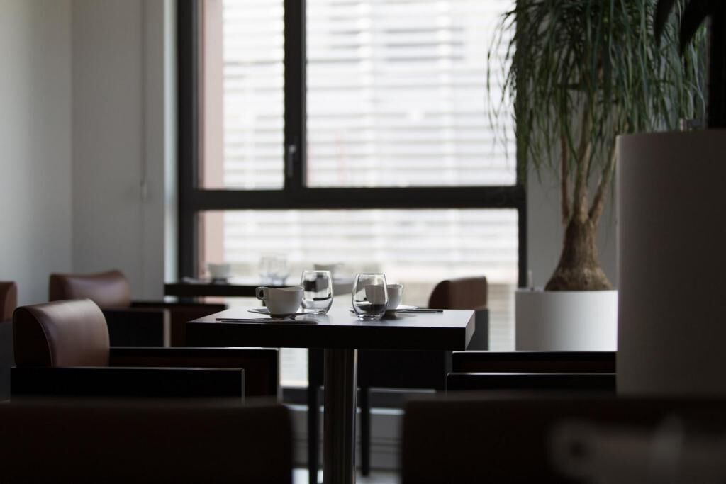 Een moderne eetruimte met een tafel voor twee personen, een potplant en een groot raam met jaloezieën.