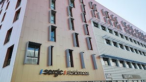 En modern byggnad med en skylt som visar texten ”Sergic Residences”, med en blandning av rosa och beige fasader, många fönster och en tydligt markerad entré.