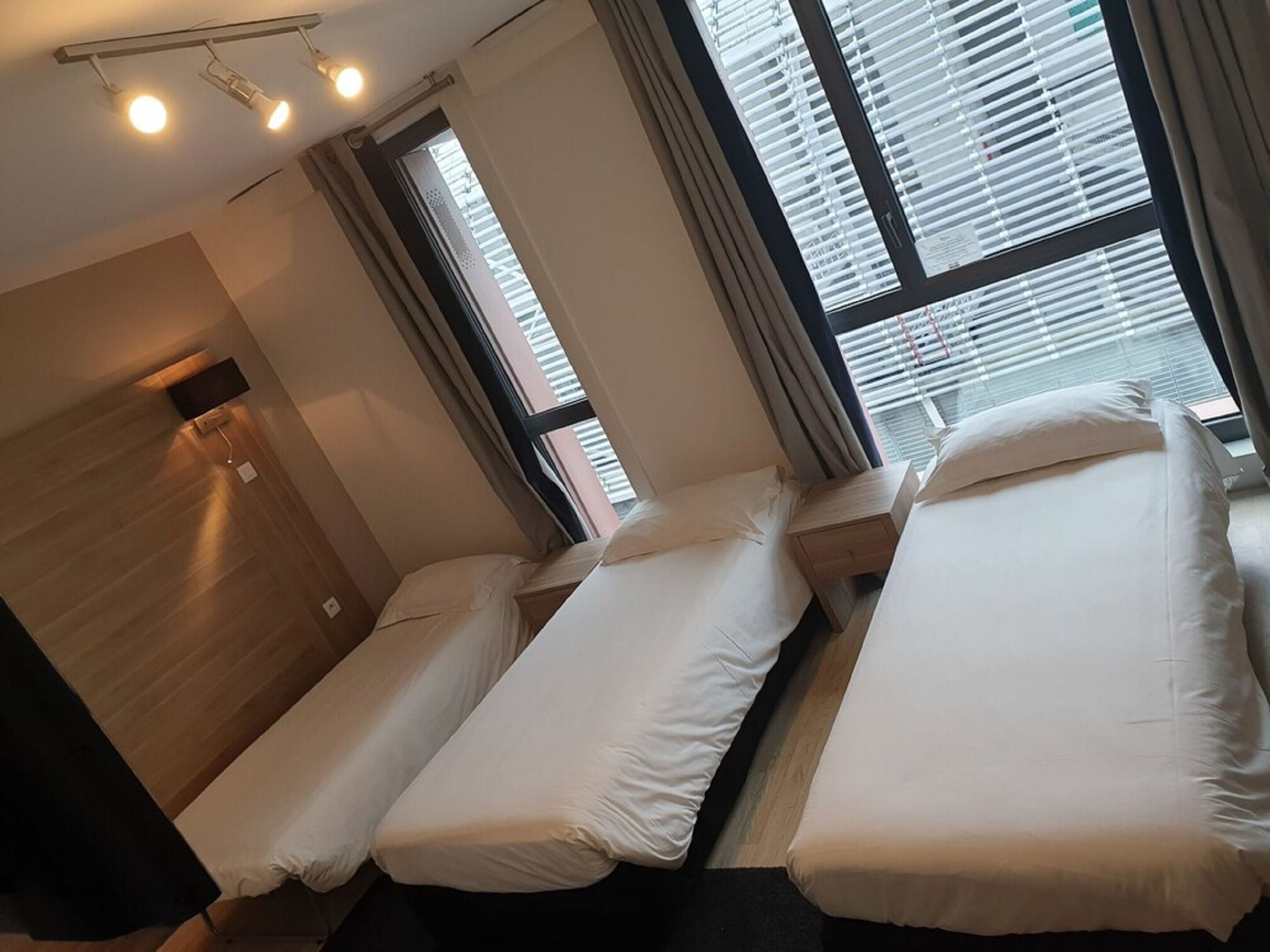 A hotel room with three single beds, a wooden headboard, a flat-screen TV, and large windows with blinds.