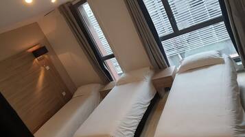 A hotel room with three single beds, a wooden headboard, a flat-screen TV, and large windows with blinds.