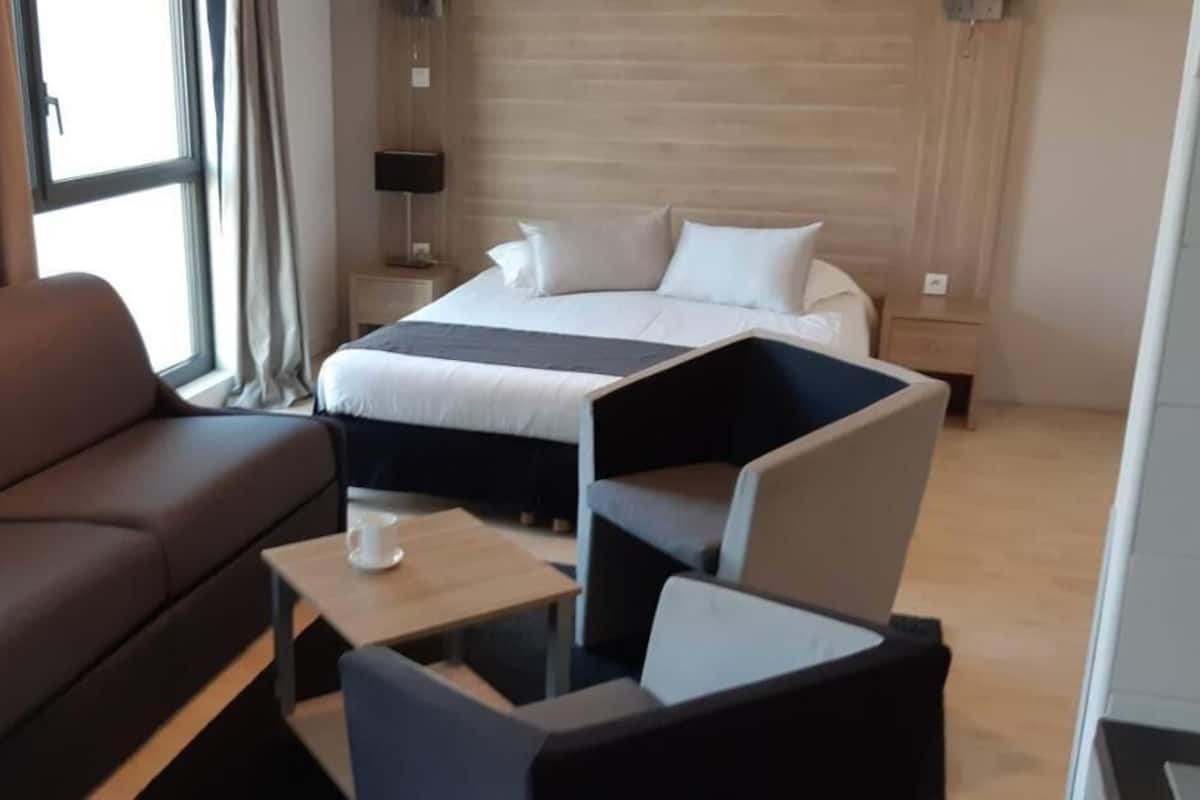 A hotel room with a brown sofa, a small table with a cup, a bed with white linens, and a nightstand with a lamp.