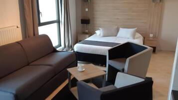 Habitación de hotel con un sofá marrón, una mesita con una taza, una cama con sábanas blancas y una mesita de noche con una lámpara.