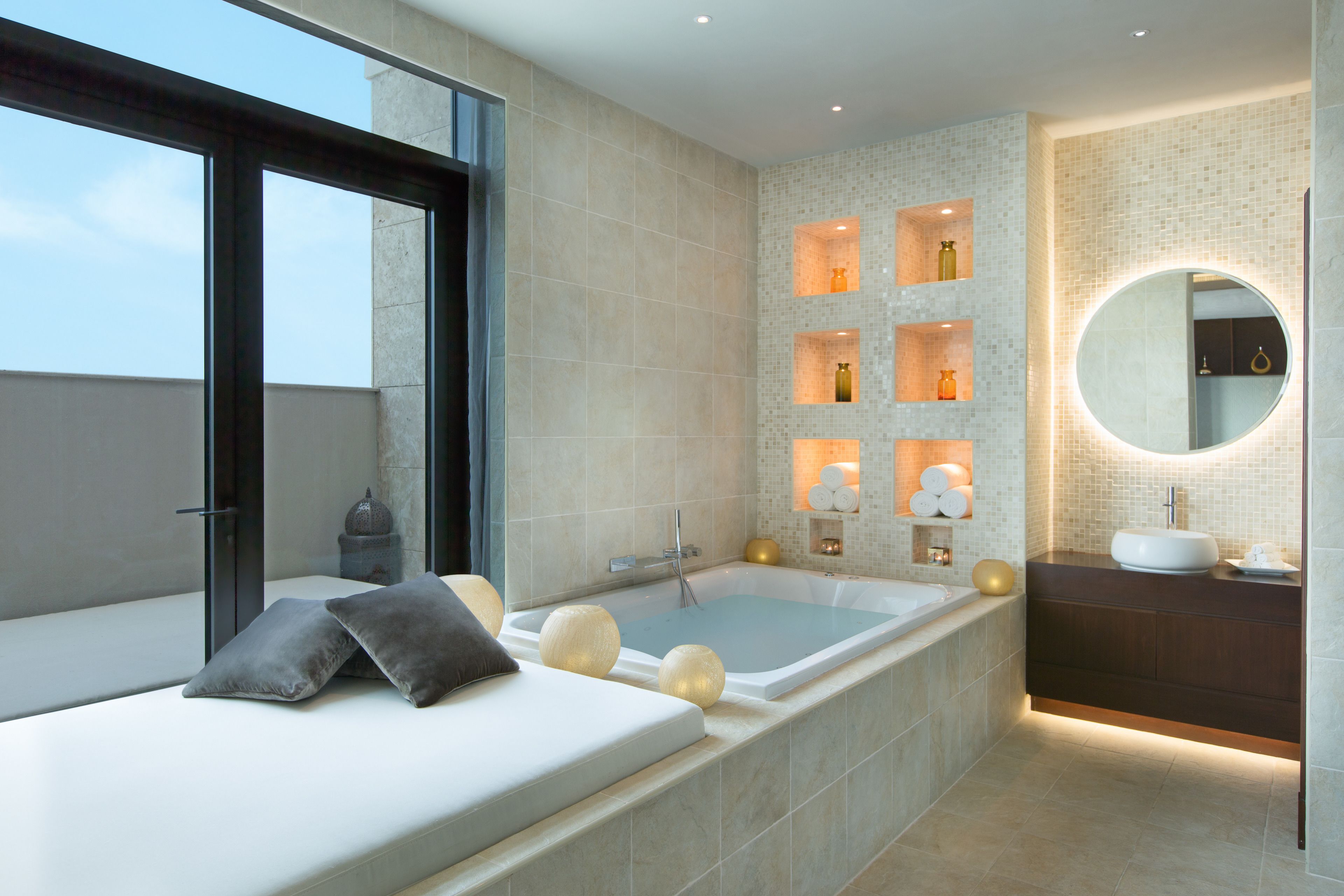 A modern bathroom with a large bathtub, a round mirror, and a built-in shelf with decorative items.
