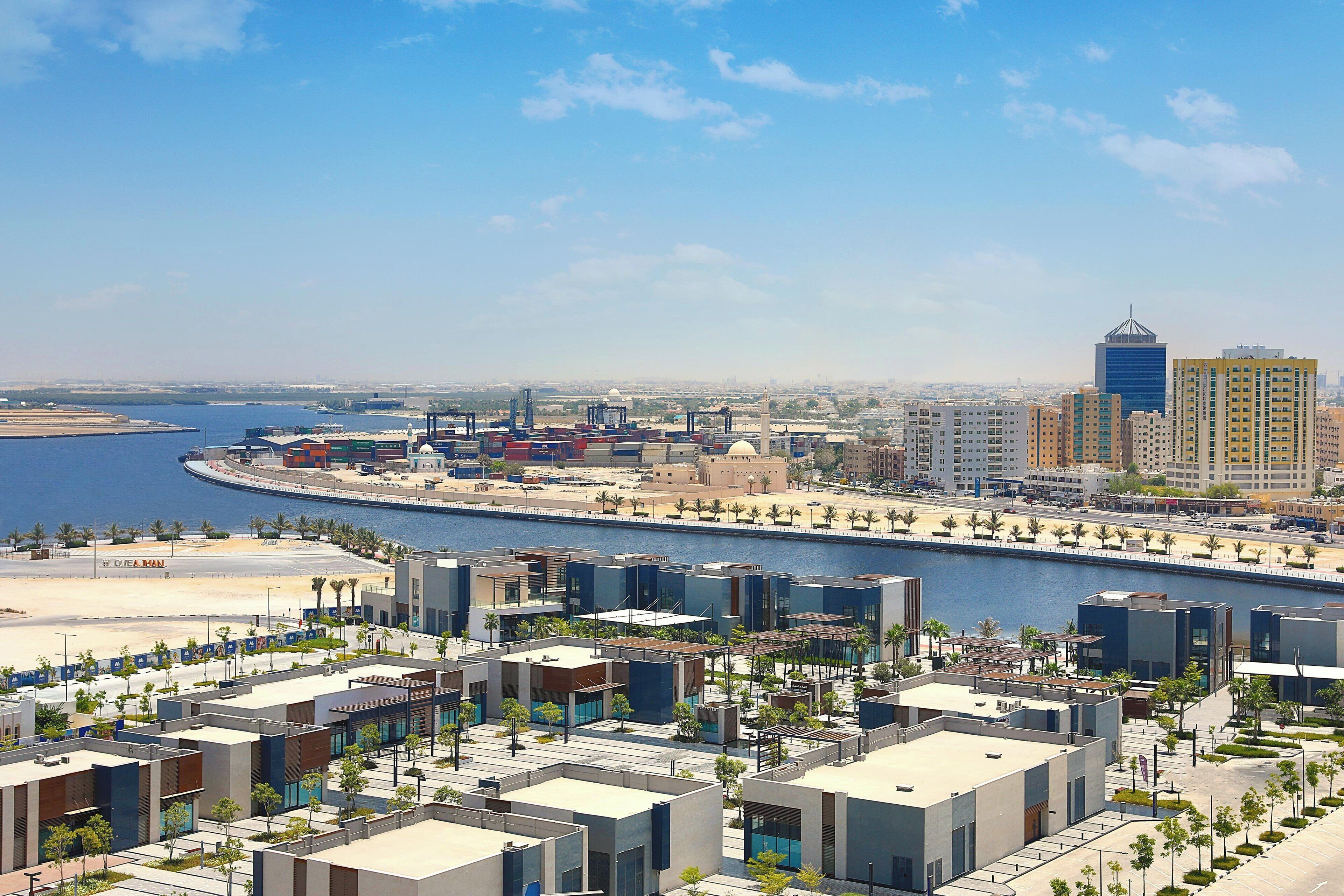 A modern urban area with a waterfront, featuring contemporary buildings, a marina, and a promenade lined with palm trees.