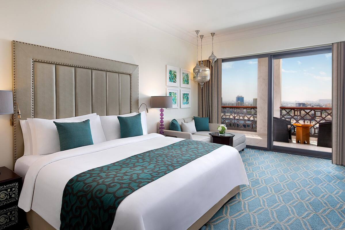 Ett hotellrum med en stor säng, en soffa, en balkong med utsikt och en stadsbild som syns genom fönstren.