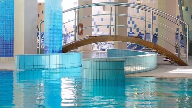 An indoor swimming pool with clear water, a wooden arch bridge, and blue chairs around the pool area. - Ramada Resort by Wyndham Kranjska Gora (Kranjska Gora)