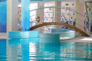 Una piscina interior con agua cristalina, un puente arqueado de madera y sillas azules alrededor de la zona de la piscina.