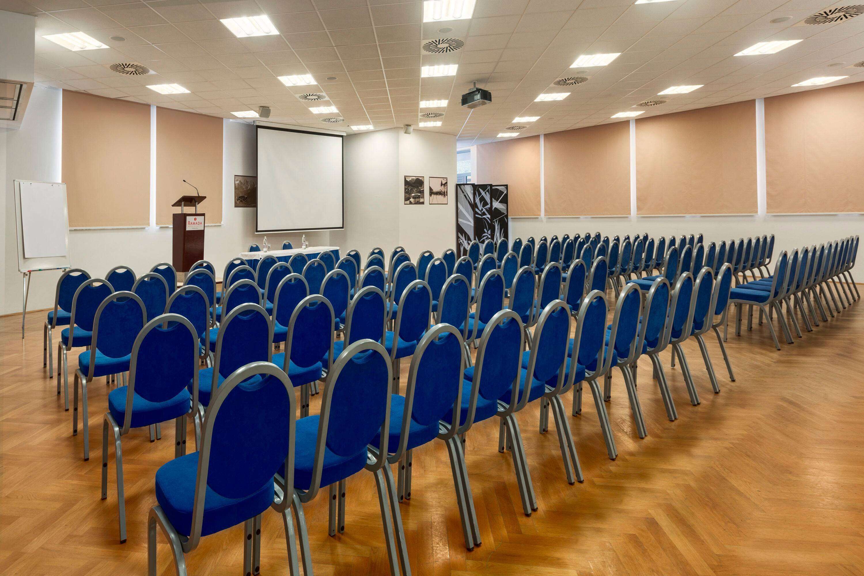 Une salle de conférence avec des rangées de chaises bleues, un pupitre, un écran de projection et un tableau blanc.