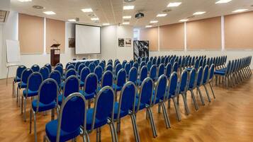 Une salle de conférence avec des rangées de chaises bleues, un pupitre, un écran de projection et un tableau blanc.
