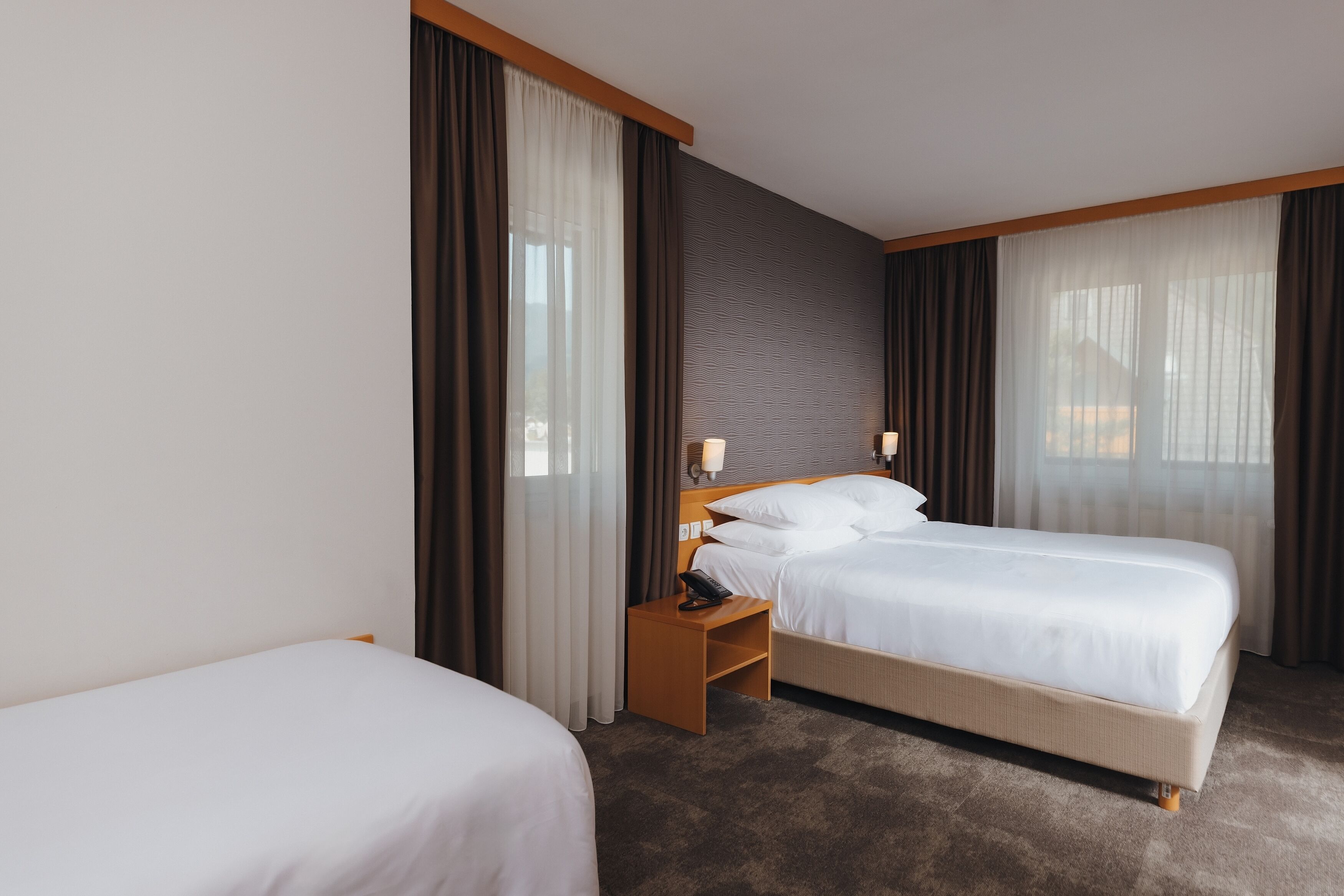 Habitación de hotel con dos camas, una mesita de noche de madera, una lámpara y vistas al exterior a través de amplias ventanas.