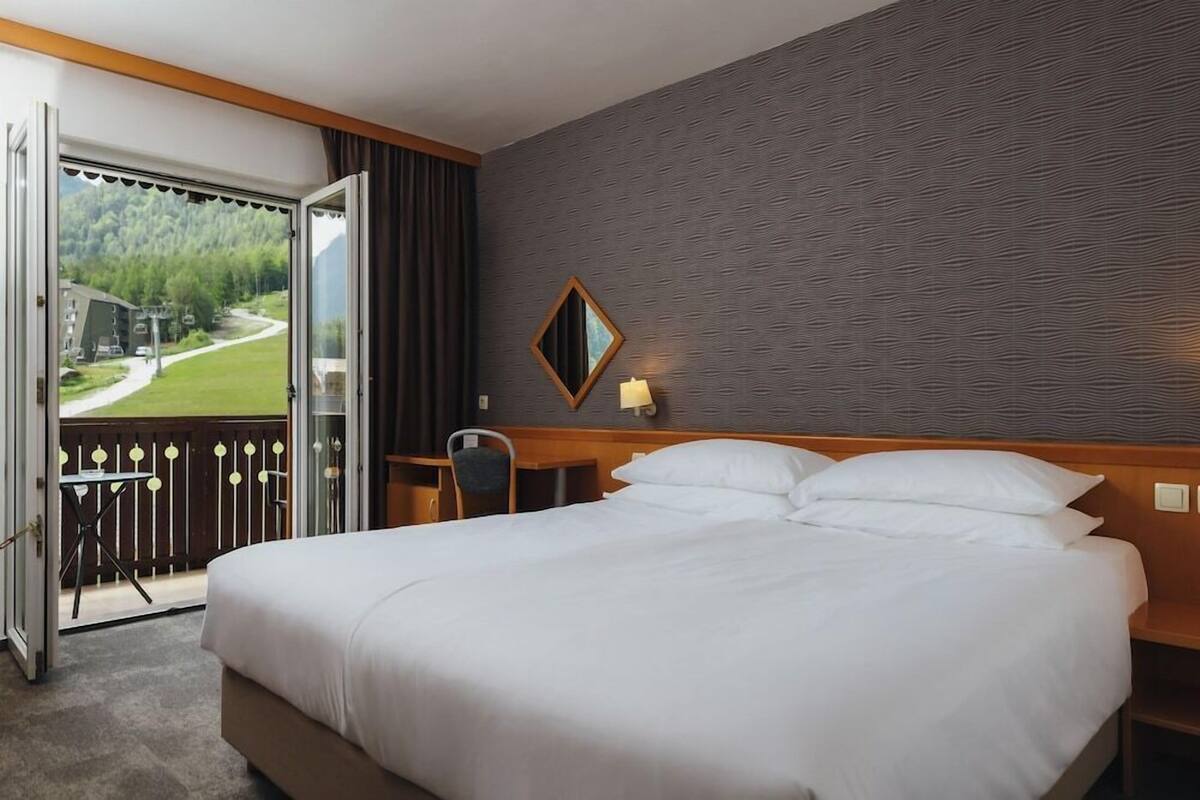 Een hotelkamer met een groot bed, een bureau, een stoel en uitzicht op een groen landschap door een geopende deur.