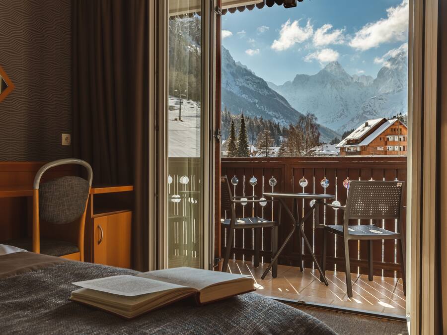 A hotel room with a bed, a desk, a chair, and a balcony with a view of mountains.