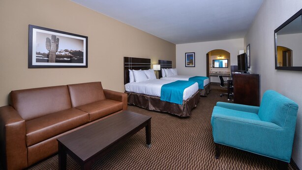 A hotel room with a brown leather sofa, a turquoise armchair, a wooden coffee table, a bed with white linens and a blue accent, a flat-screen TV, a desk with a chair, and a framed picture on the wall. - Americas Best Value Inn & Suites Cuero (Cuero)
