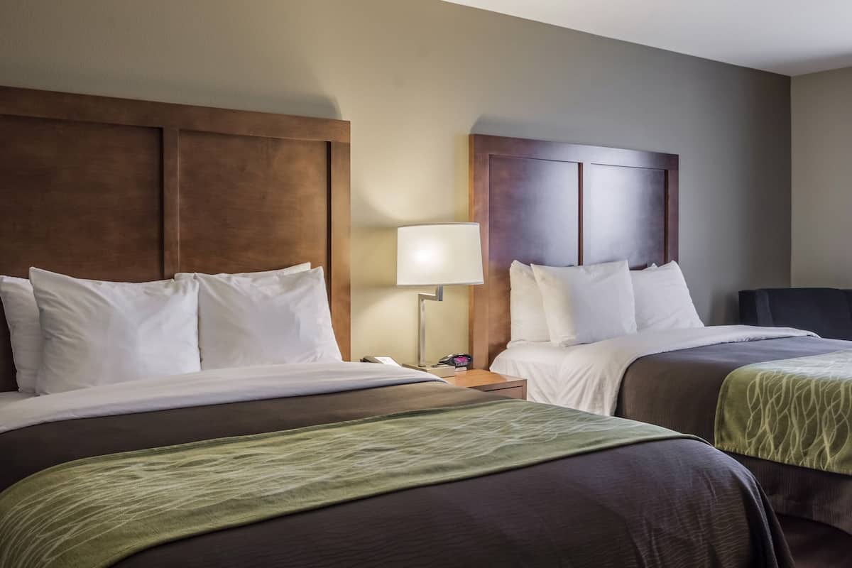 A hotel room with two beds, each with white linens and a wooden headboard. There is a bedside table with a lamp and a clock.