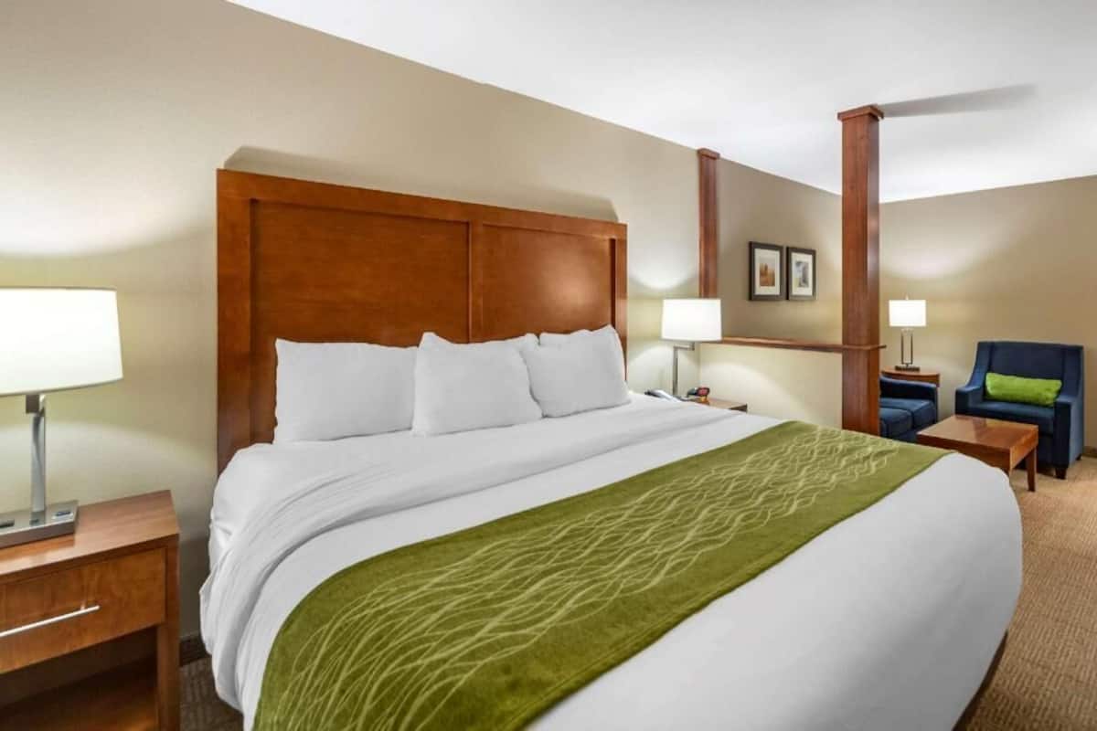 A hotel room with a large bed, a wooden headboard, two bedside tables with lamps, a chair, and a small table.