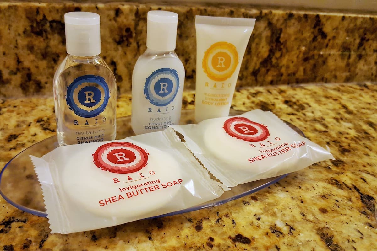Three bottles of RAIO hair care products and two packets of RAIO Shea Butter Soap on a granite countertop.