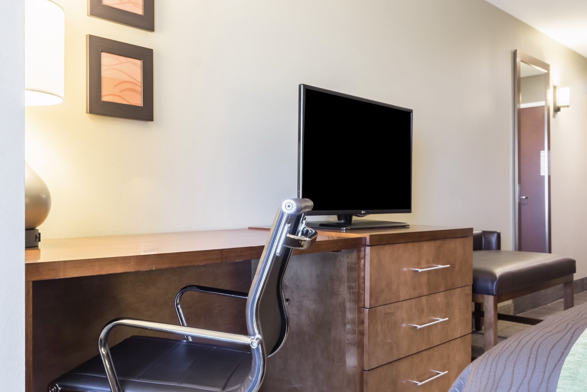 A hotel room with a wooden desk, a black chair, a television, a dresser, and a bench.