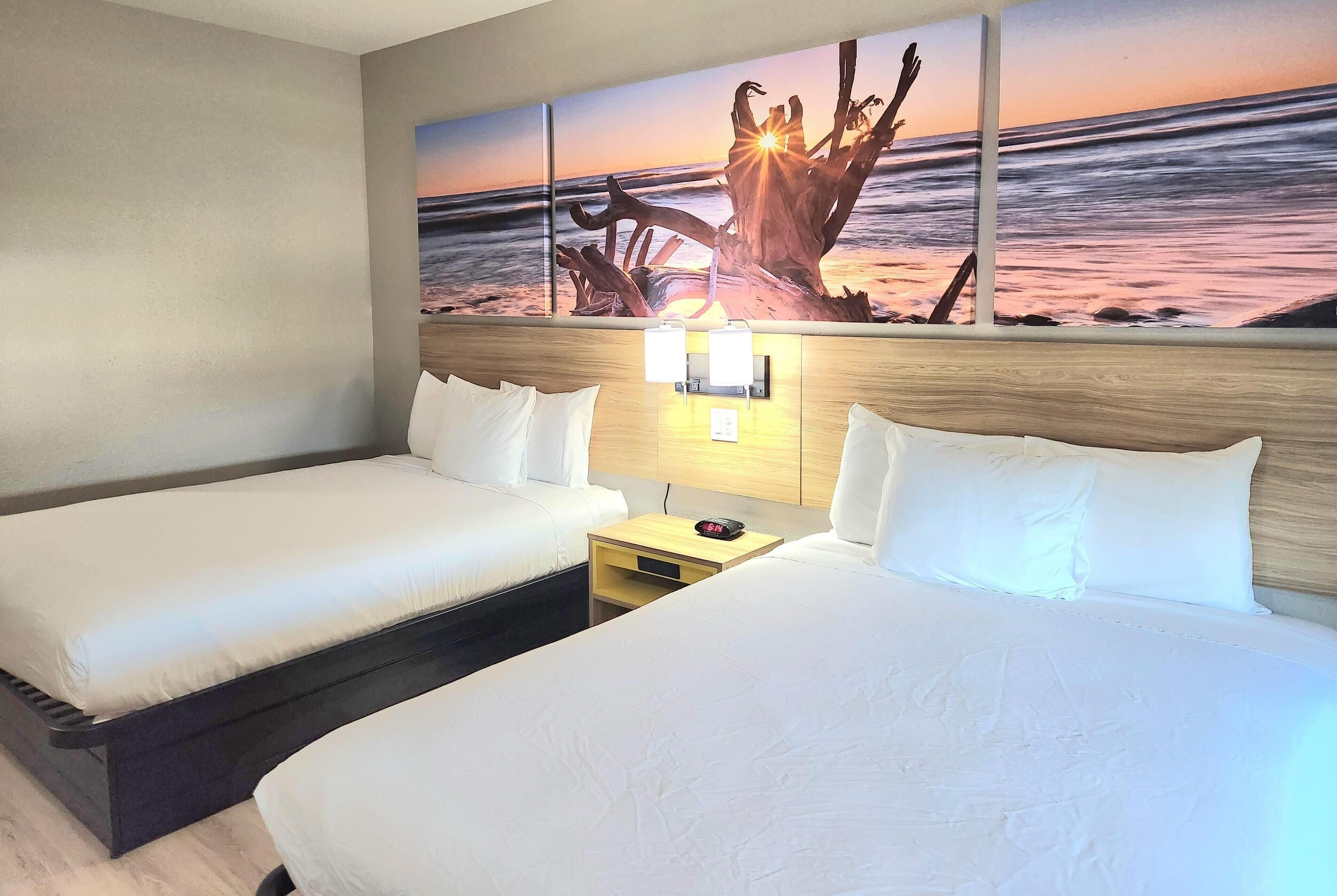 Habitación de hotel con dos camas, una mesita de noche, una lámpara de pared y un cuadro de una puesta de sol sobre el océano.