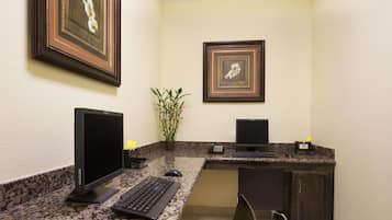 A hotel room with a desk, two monitors, a keyboard, a mouse, a chair, a telephone, and framed pictures on the wall.