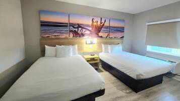 A hotel room with two beds, a large wall mural of a sunset, a bedside table with a lamp, and a window with blinds.