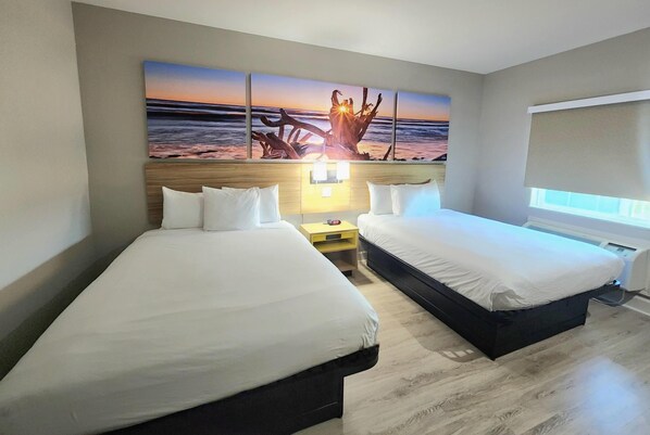 A hotel room with two beds, a large wall mural of a sunset, a bedside table with a lamp, and a window with blinds.