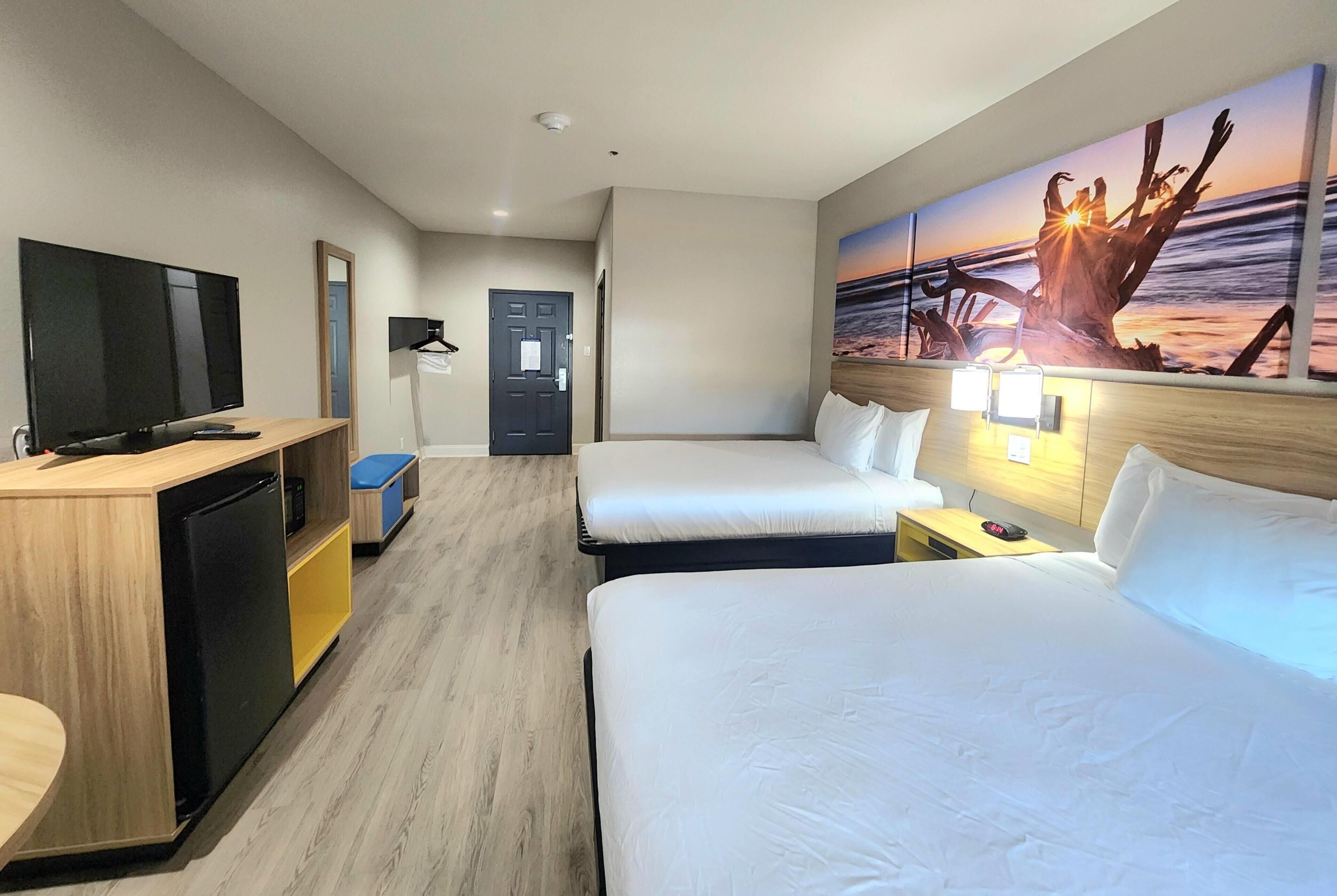 A hotel room with two beds, a desk with a TV, a small kitchenette, and a painting of a sunset over the ocean.