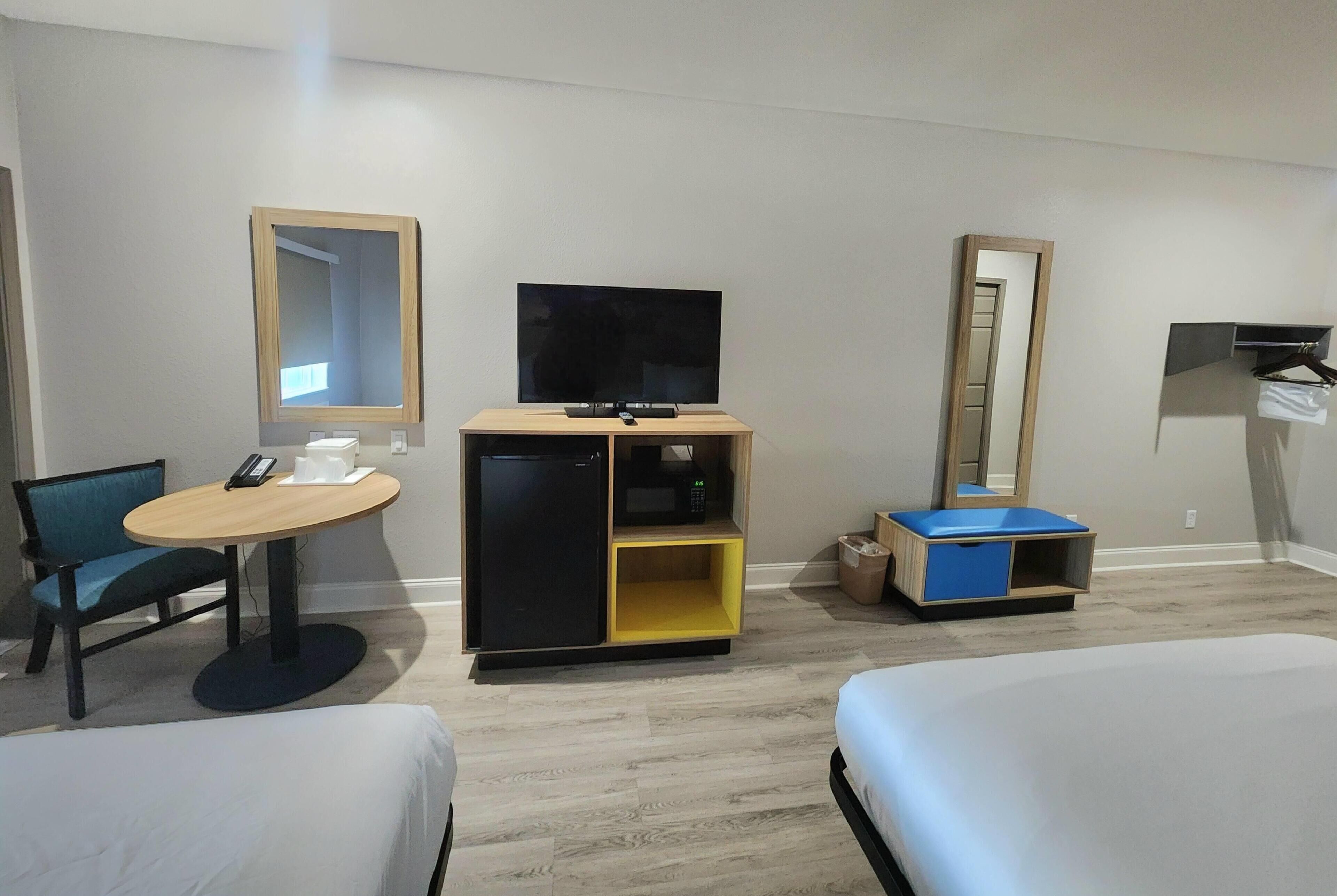 Habitación de hotel con cama, escritorio con silla, televisión, refrigerador pequeño y espejo.