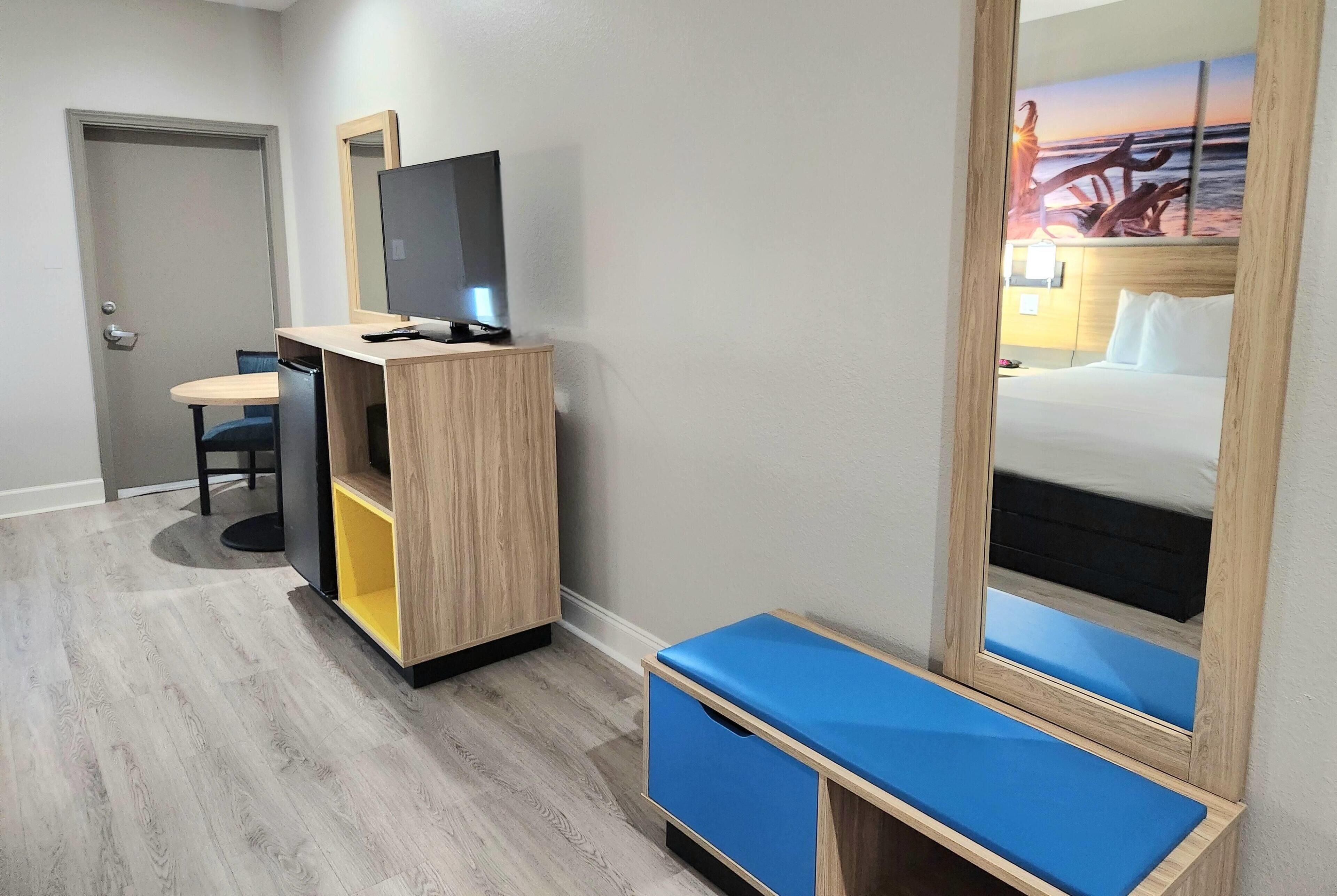 A hotel room with a wooden desk, a television, a mirror, and a bed with a blue headboard.