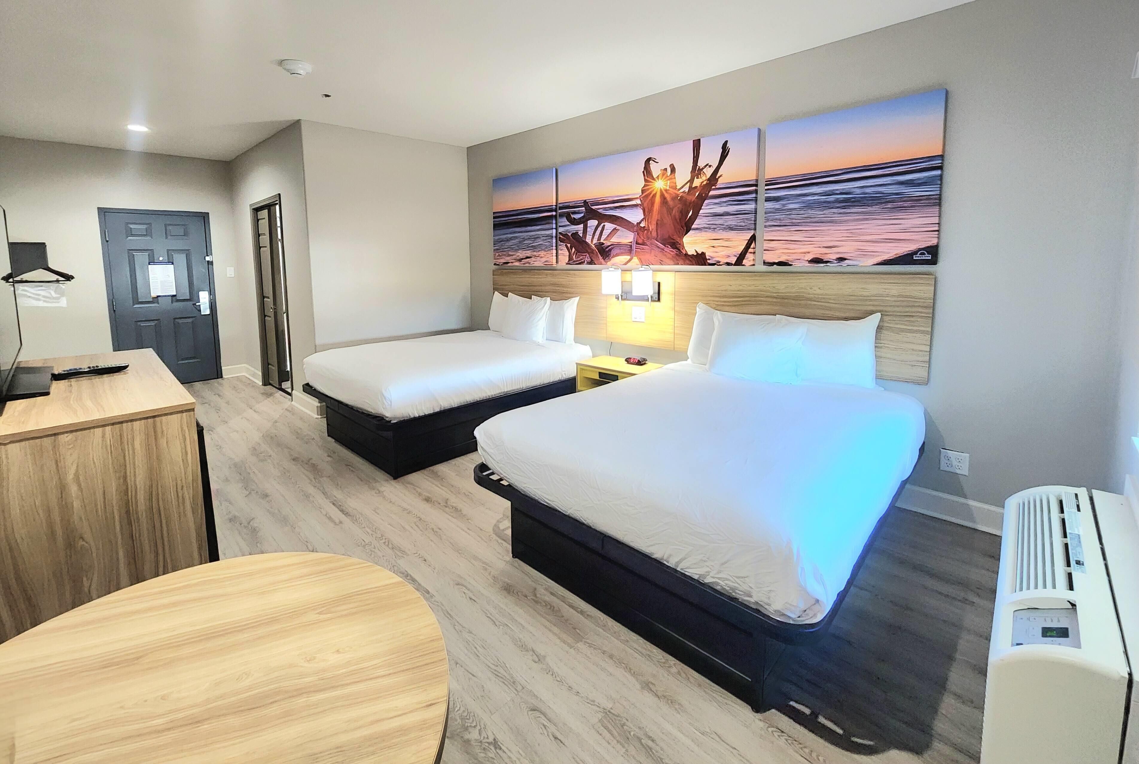 A hotel room with two beds, a desk, a chair, and a painting of a sunset.