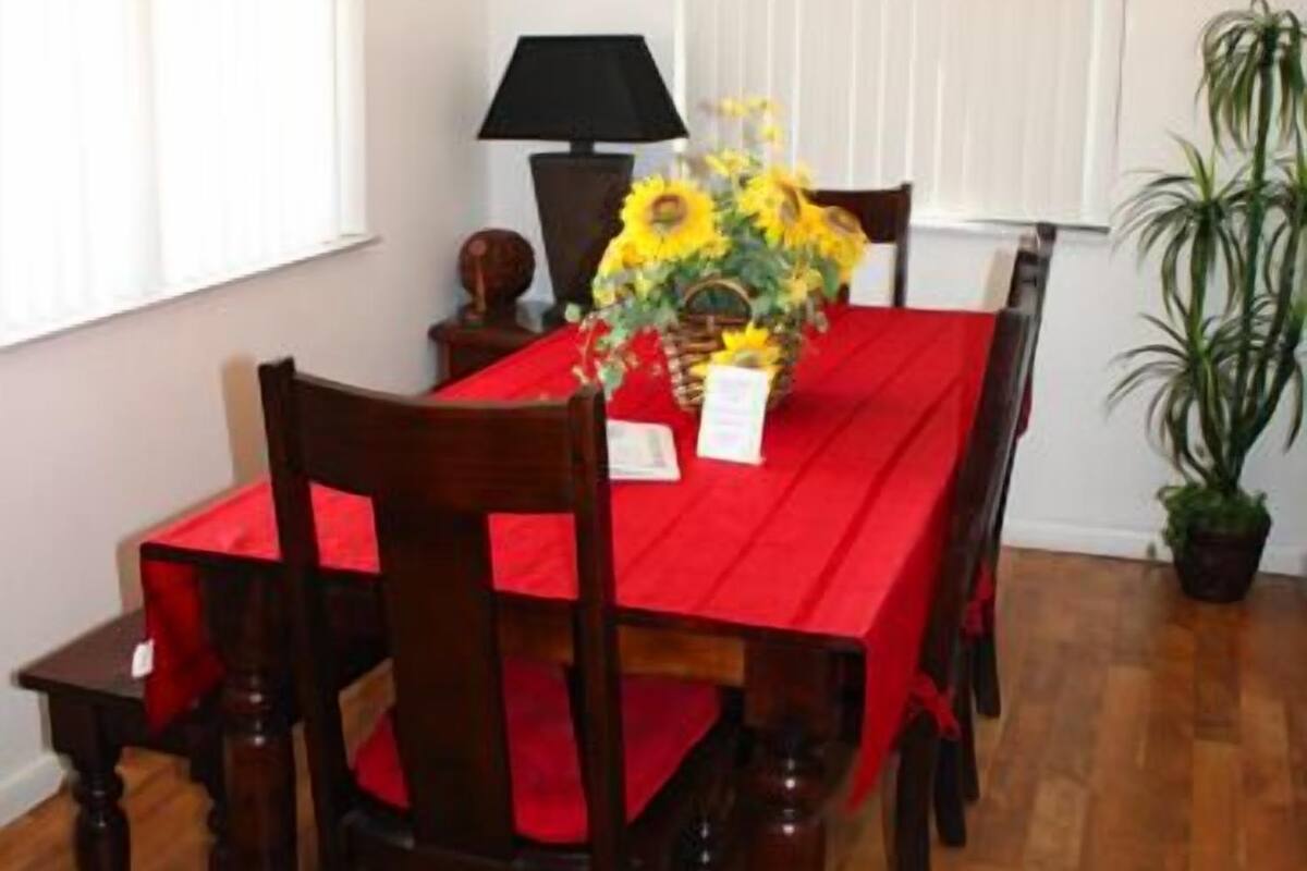 Un coin repas avec une nappe rouge, des chaises en bois, un vase rempli de tournesols, un luminaire suspendu et une plante en pot.