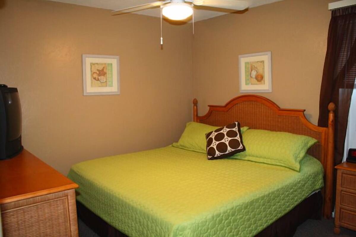 Un lit simple avec une couette verte, une tête de lit en bois et un ventilateur de plafond.