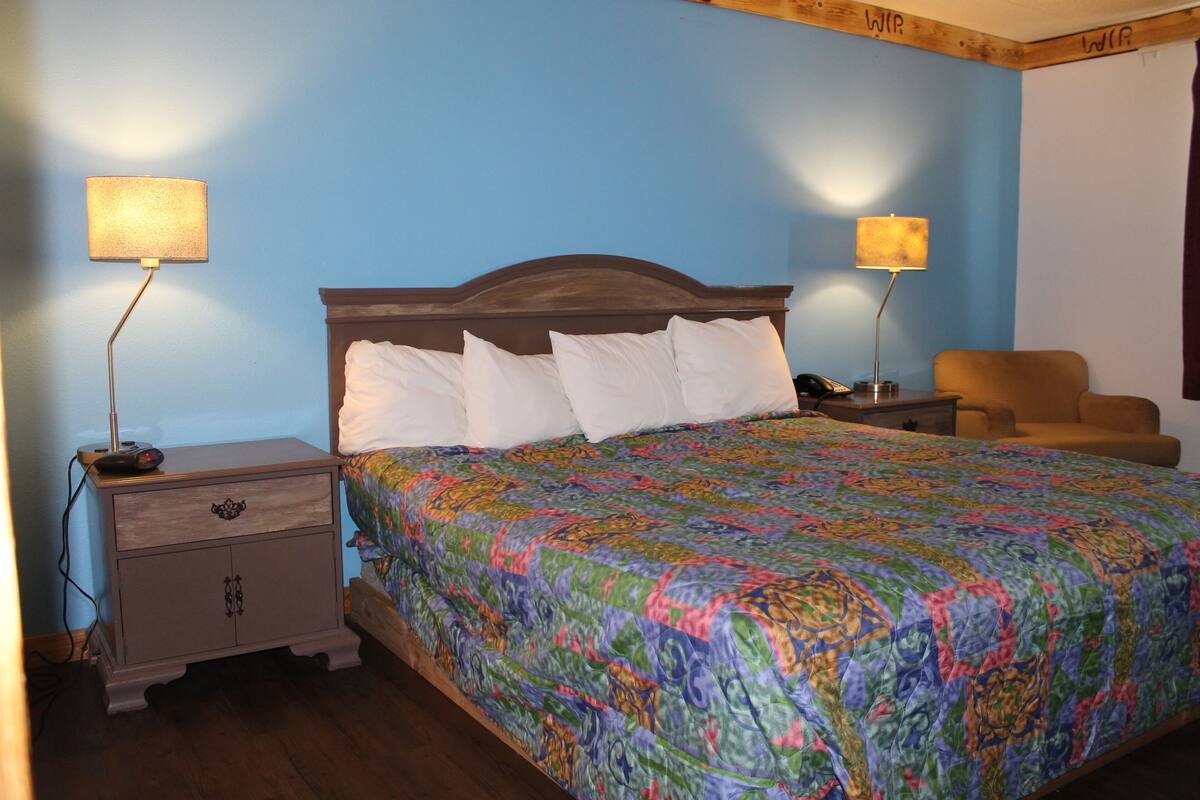 Ein Schlafzimmer mit einem hölzernen Kopfteil, einer farbenfrohen Stegdecke, zwei Nachttischlampen, einem Nachttisch und einem Sessel.