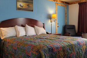 Habitación de hotel con una cama grande, un cabecero de madera, una mesita de noche con lámpara, una silla y una puerta con cortinas.