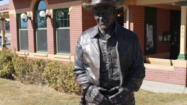 A statue of a man in a hat and vest standing in front of a building with a sign that reads 'Visitor Information.' - Wyatt Earp Hotel (Dodge City)