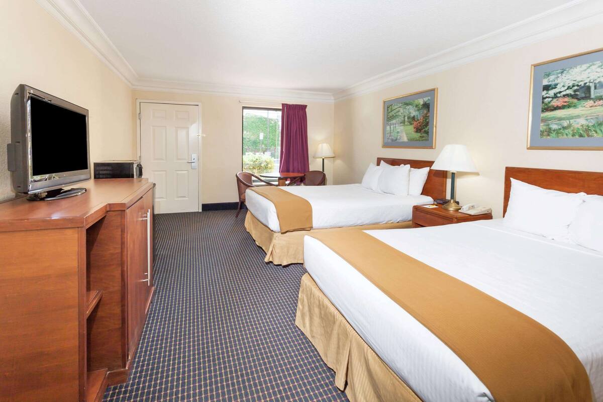 A hotel room with two beds, a television, a desk, and a window with a view of greenery.