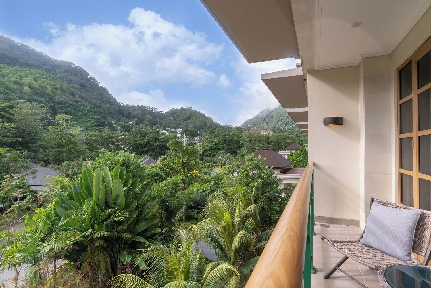 Een balkon met een rieten stoel en een uitzicht op weelderige begroeiing en huizen op de achtergrond.