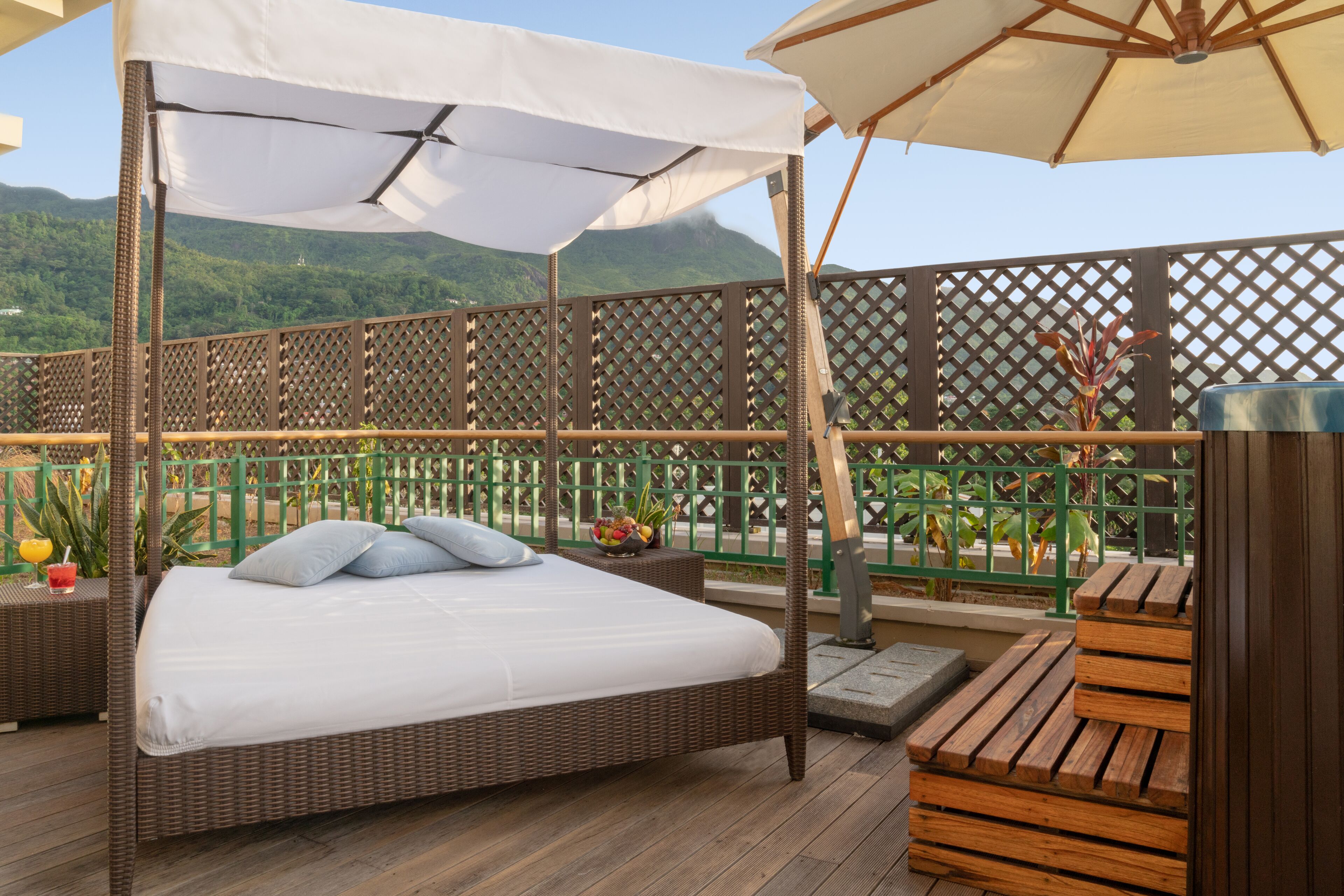 Uma varanda com cama de vime, assentos de madeira e vista para colinas verdes.