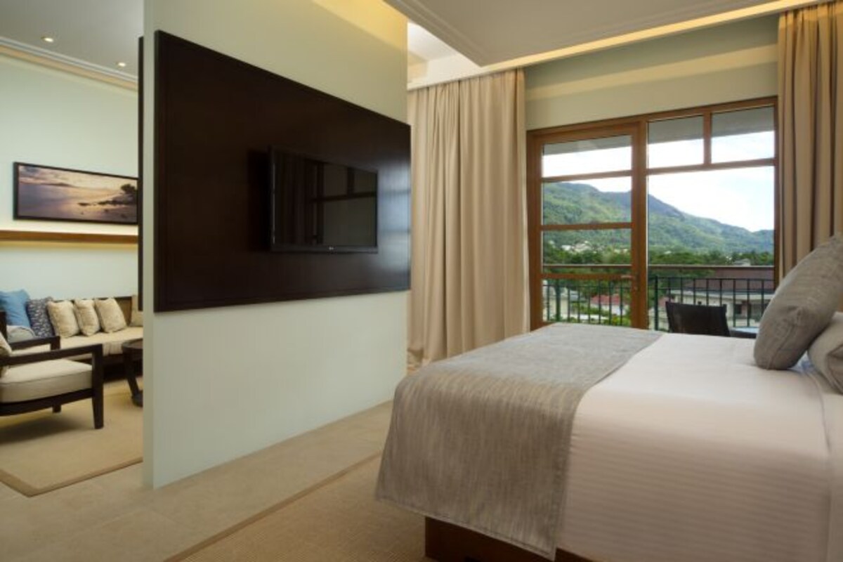 A hotel room with a bed, a TV mounted on the wall, a sofa, and a balcony with a view of the mountains.