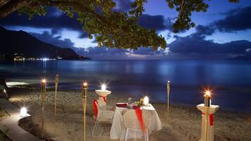 A beachside dining setup with tables and chairs, candles, and a view of the sea and mountains at night.