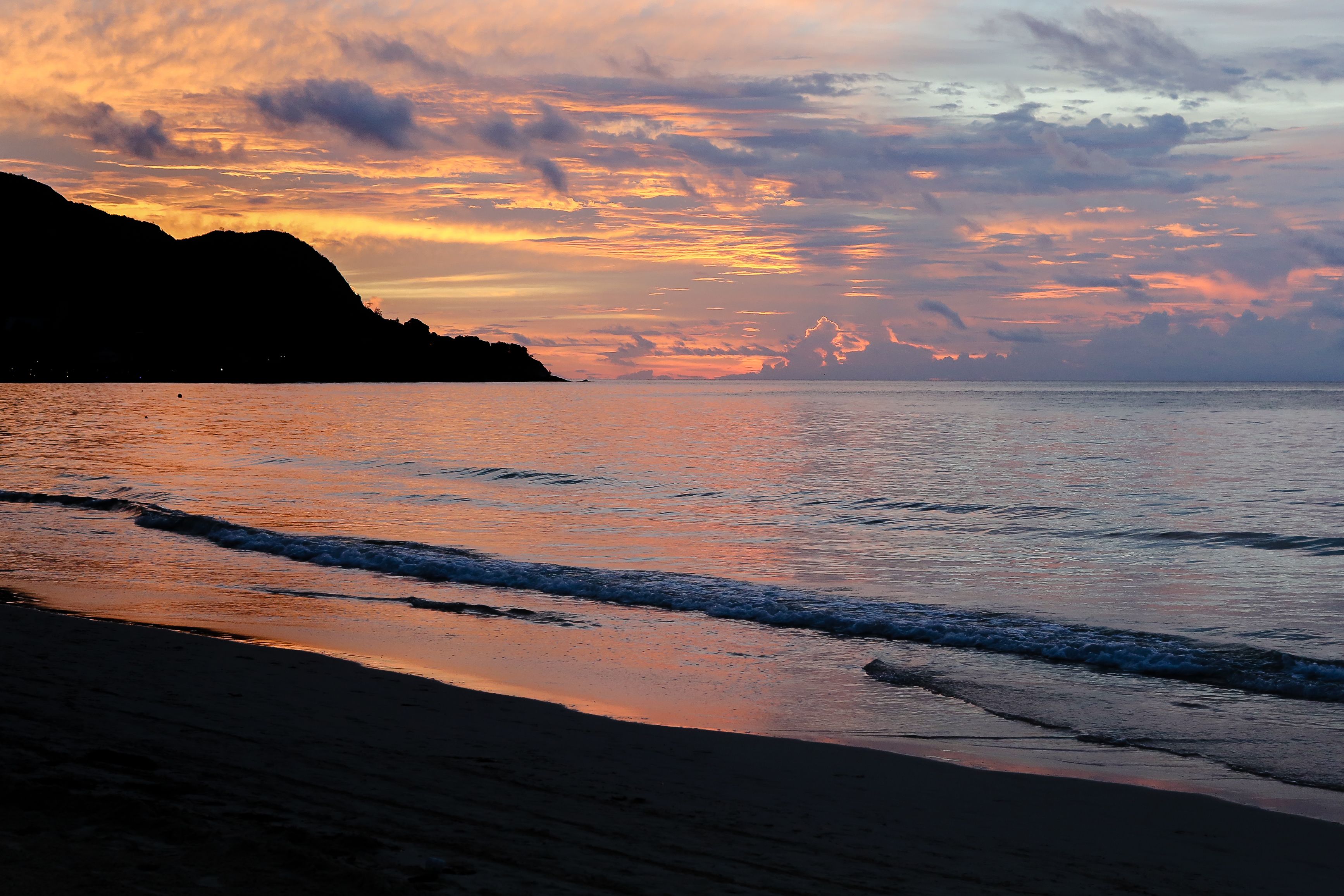 A beach with waves at sunset, a silhouette of a mountain, and a clear sky.